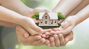 Hands holding a miniature house surrounded by greenery, symbolizing community support in selling inherited or distressed properties.