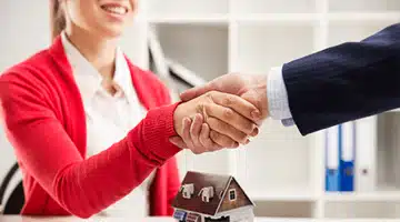Woman in red cardigan shaking hands with a man in a suit, with a small model house on the table, symbolizing a successful real estate transaction.