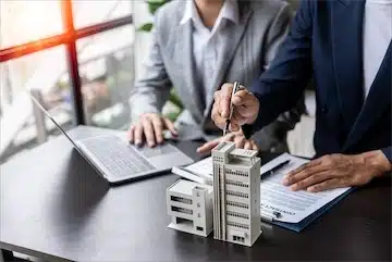 Business professionals discussing commercial property sale with a miniature building model and documents on a desk.