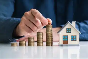 Hand placing a coin on a stack of coins beside a miniature house, symbolizing investment in rental properties and cash offers from ROI National.