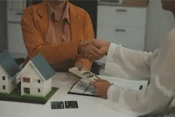 Handshake between individuals at a table with miniature houses and documents, representing a smooth real estate transaction for selling homes.